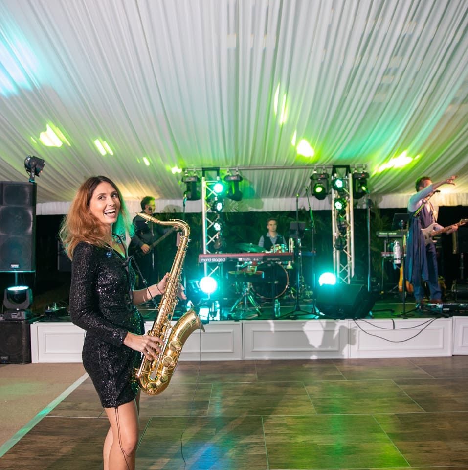 Amy Cyr laughing with her saxophone in front of a full band under an event tent